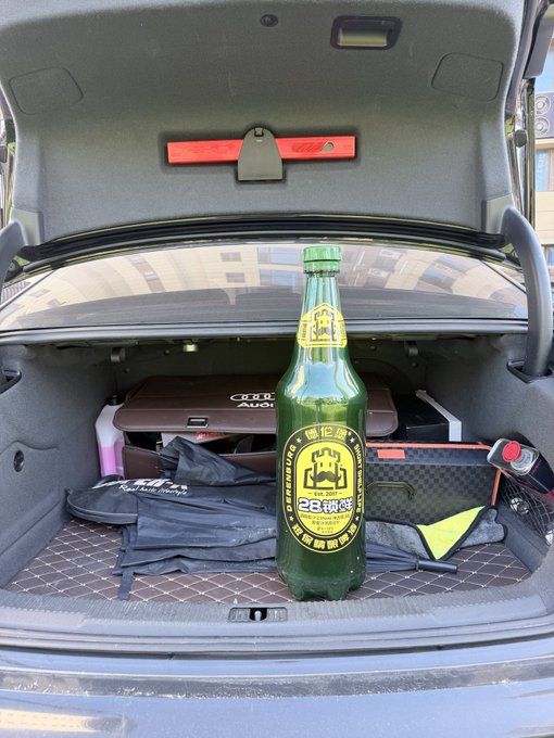 A large green bottle of 'Tsingtao Beer' (青島啤酒, 28 copper medal edition) sitting in the open trunk of an Audi, alongside an Audi-branded box and accessories.