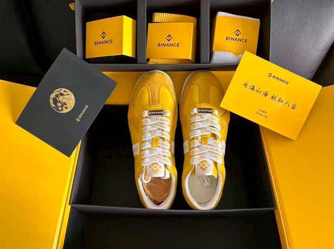 A photograph of a Binance-branded yellow and white sneaker gift box set, including sneakers, Binance packaging, a Bitcoin-themed card, and a yellow envelope with Chinese calligraphy.