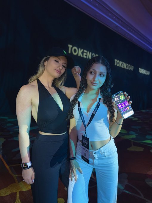 Two women pose together at a TOKEN2049 event, one wearing a black halter outfit and cap, the other in a white top and pants showing a colorful game-like display on her phone screen.