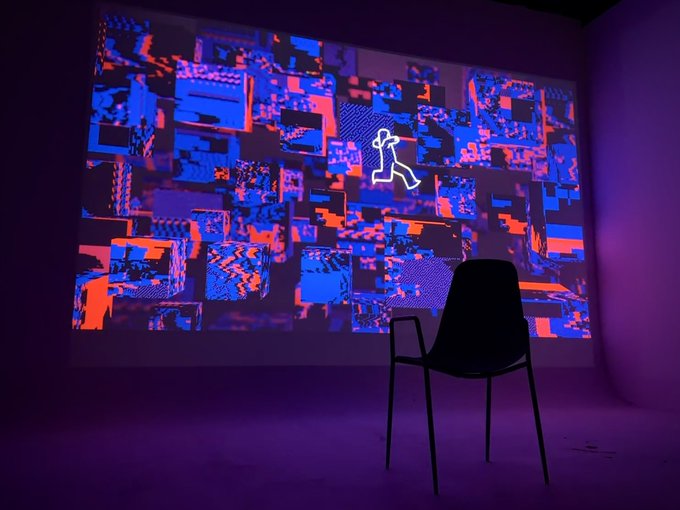 A dimly lit room with a black chair facing a large wall projection displaying abstract red and blue pixelated glitch art featuring a walking figure.