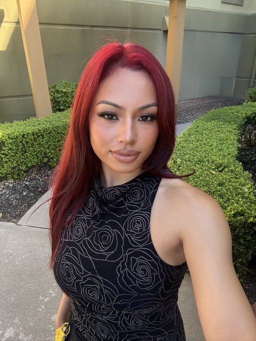 A woman with long red hair and a black sleeveless top with a rose pattern takes a selfie outdoors near bushes and a building.