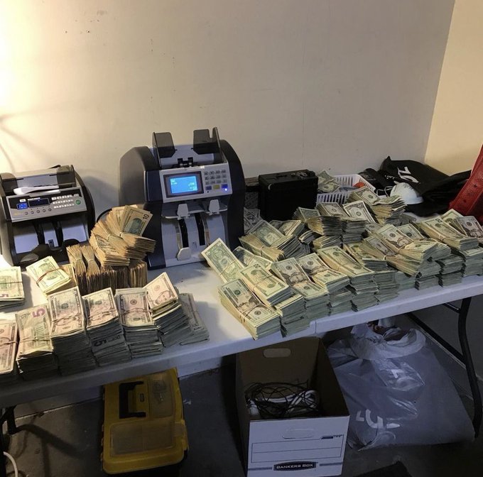 A table covered in bundles of US dollar bills with two money-counting machines and a yellow toolbox, with white plastic bags and a Bankers Box on the floor below.