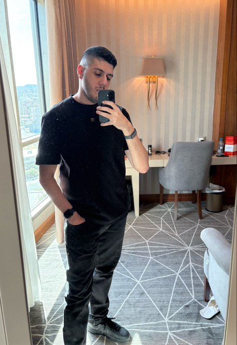 A man in a black t-shirt and black pants takes a mirror selfie in a hotel room, holding a phone to take the photo while standing near a window with a view outside.