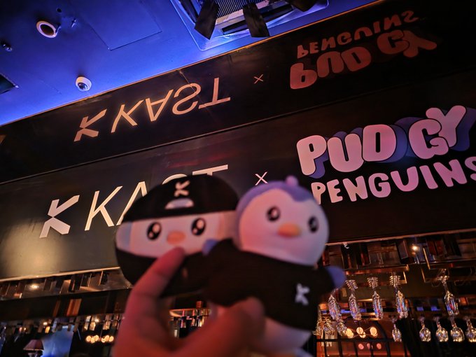 A hand poses two "Pudgy Penguins" plush toys in front of a dimly lit bar with illuminated signage reading "KKKK," "TSAXK," "GO PENGUINS," and "NO GO HOME," capturing the lively atmosphere of a KAST x Pudgy Penguins collaboration event.