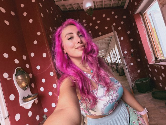 A selfie of a woman with vibrant pink hair wearing a patterned top, standing in a red room decorated with white polka dots.