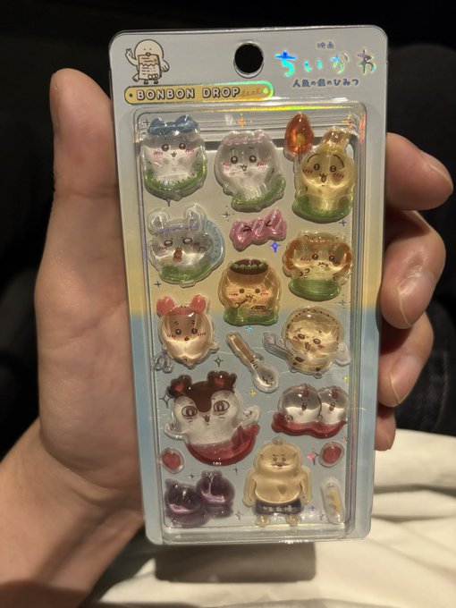 A hand holds up a packaged set of Chiikawa "BONBON DROP" stickers featuring various characters, humorously tied to a father who was guilted into watching the Chiikawa movie by his family, only to realize their true motive was getting these collectible stickers.