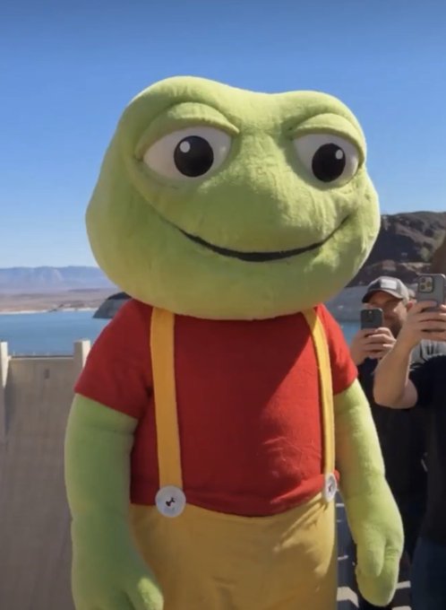 A photo of a person in a Pepe the Frog mascot costume wearing a red shirt and yellow overalls, standing outdoors near what appears to be a body of water, with people taking photos in the background.