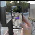 A person holding a smartphone taking a photo of a purple-haired character illustration on a street.