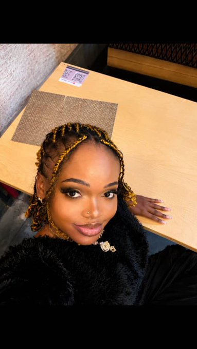 A selfie of a young woman with braided hair wearing a fuzzy black jacket, taken indoors near a wooden table.