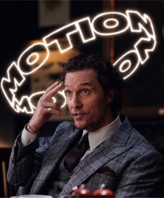 A man in a gray plaid suit gestures with his hand near his temple in front of a neon "MOTION" sign, wearing a bracelet and ring.