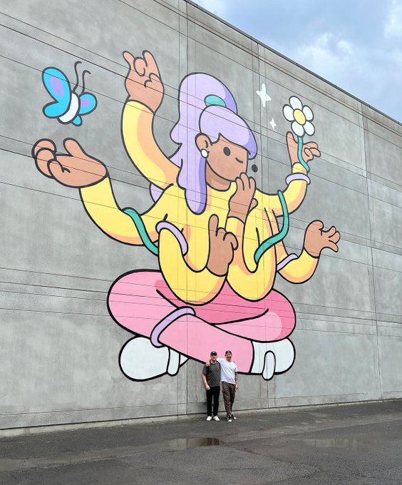 Two people stand at the base of a large mural on a concrete wall depicting a multi-armed cartoon figure in a yellow sweater and pink pants holding a flower, with a blue butterfly nearby.