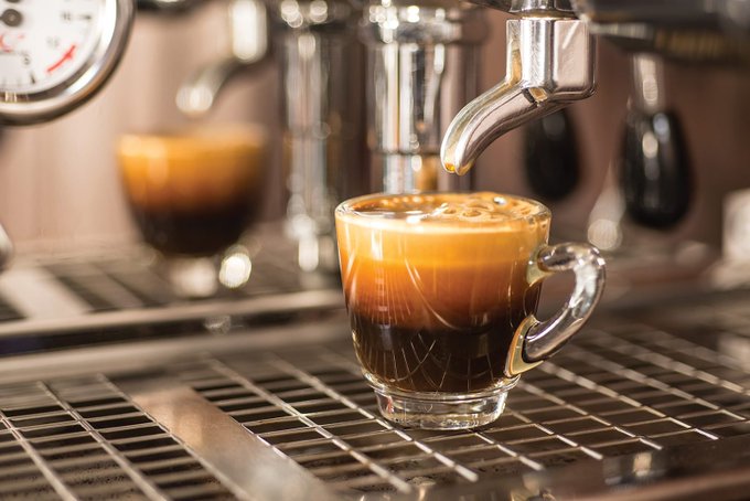 A freshly pulled shot of espresso fills a small glass cup on a professional espresso machine's drip tray, with a pressure gauge visible in the upper left corner. The rich, dark espresso matches the post's warm greeting inviting followers to enjoy coffee together.
