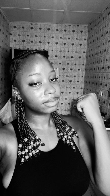 A black and white selfie of a woman with long beaded braids wearing a black tank top, posed in front of patterned wallpaper.