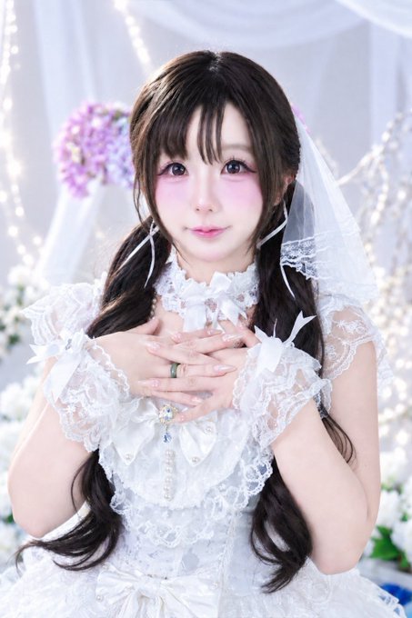 A young woman with long brown hair wearing an ornate white lace dress and veil, posing with her hands crossed over her chest in front of a floral backdrop.
