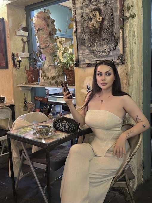 A woman with dark hair and tattoos sits at a small café table in a beige strapless dress, holding a phone with a stylish handbag on the table.