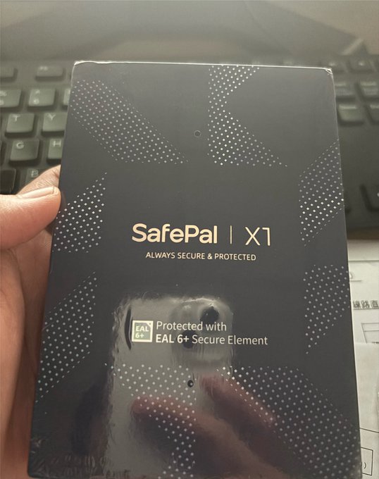 A hand holding a black SafePal X1 hardware wallet box with the tagline 'Always Secure & Protected' and a note indicating it is protected with EAL 6+ Secure Element.