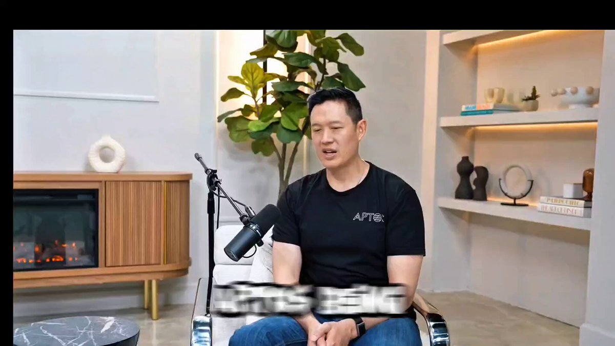 The image shows a man sitting in an office chair, wearing a black shirt with white text that says "aptos" on it. The setting appears to be a conference room or office space, as there are several books visible around him. The background features a fireplace and a potted plant, adding to the overall atmosphere of the scene.