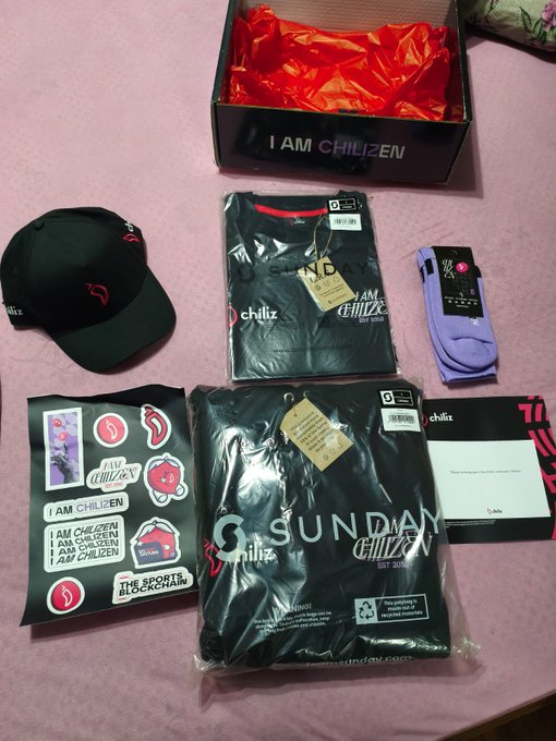 The image shows a collection of items, including clothing and accessories, arranged on a pink surface. The main focus is a black t-shirt with white text that reads "I am Chiliz", along with other clothing items such as a hat, socks, and a shirt. There are also several stickers scattered around the scene.

The post features an image of the items displayed in the box, which includes a black t-shirt, a hat, and some accessories like socks. The text on the post reads "I am Chiliz @alex_dreyfus @socios".