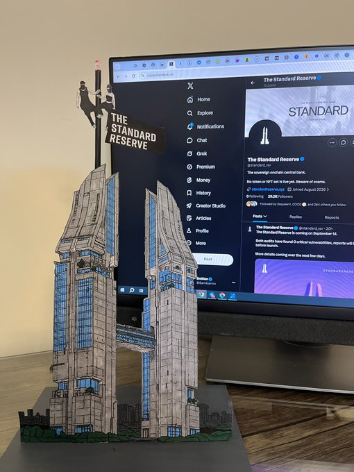 A detailed 3D-printed model of The Standard Reserve building sits in front of a computer monitor displaying the project's verified X (Twitter) profile @standard_rsv, which describes itself as a sovereign onchain central bank. The accompanying post explains how the creator was inspired to build a physical representation of the project while writing content for it, transforming a digital concept into a tangible piece.