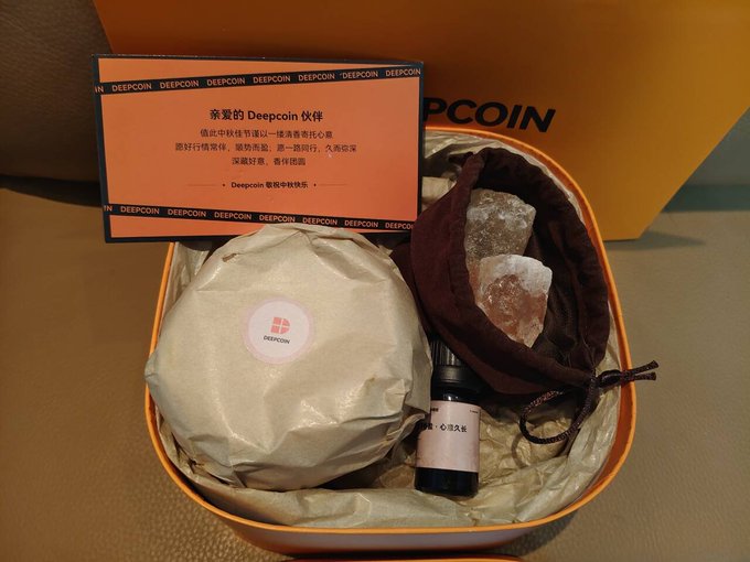 An unboxing of a Deepcoin promotional gift package containing a wrapped item, a small bottle of heart-water drops, and Himalayan salt rocks in a velvet pouch, accompanied by a card addressed to a 'Deepcoin 伙伴' in Chinese.