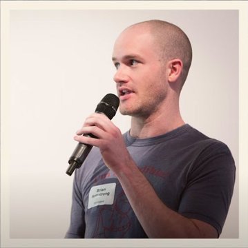 A bald man wearing a name tag labeled "Brian Armstrong" speaks into a microphone, representing the Bitcoin and crypto pioneer whose early work on mobile Bitcoin wallets preceded the founding of Coinbase. The image serves as a visual identification of Brian Armstrong, the subject of the historical milestone highlighted in the accompanying post.