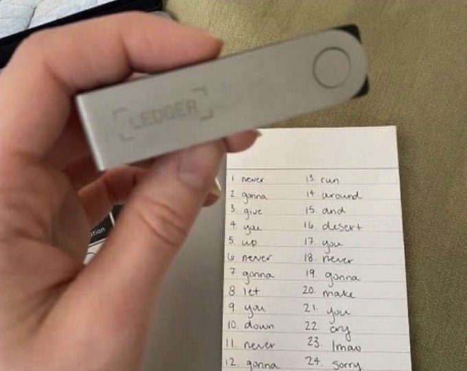 A hand holds a Ledger hardware wallet above a handwritten 24-word seed phrase recovery list (with words like "never," "gonna," "give," "you," "up"), humorously illustrating the post about securing crypto on a Ledger—where the playful "Never Gonna Give You Up" word list highlights the importance (and irony) of safely storing recovery phrases.
