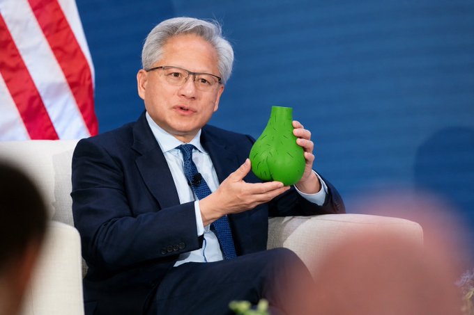 A man in a dark suit and blue tie, appearing to be Jensen Huang, sits on a white chair holding a green vase-shaped object, with an American flag visible in the background.