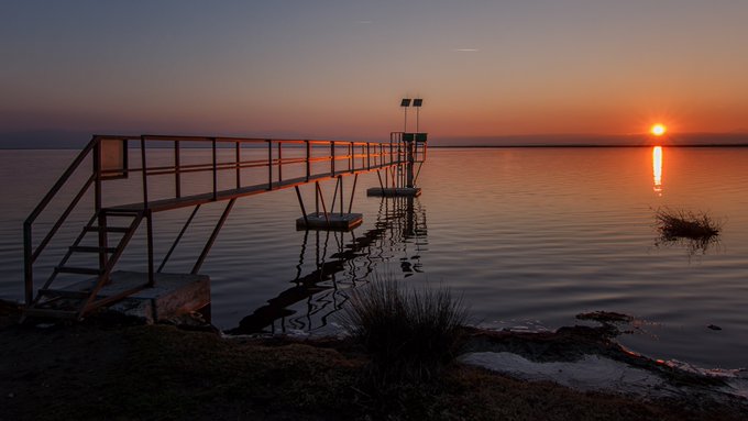 A serene sunset scene over calm water features a metal pier with stairs on the left, the sun reflecting brightly on the right, and dark silhouettes of land and vegetation.