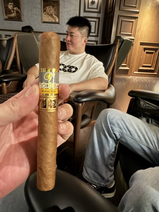 A hand holds up a Cohiba cigar (with its distinctive "Cohiba Habana, Cuba" gold and black band) in the foreground, while a man in a white graphic t-shirt relaxes in a leather chair in a wood-paneled lounge in the background—capturing the essence of the post "好茄配好" (a good cigar paired with good company) tagging @doushi0503.