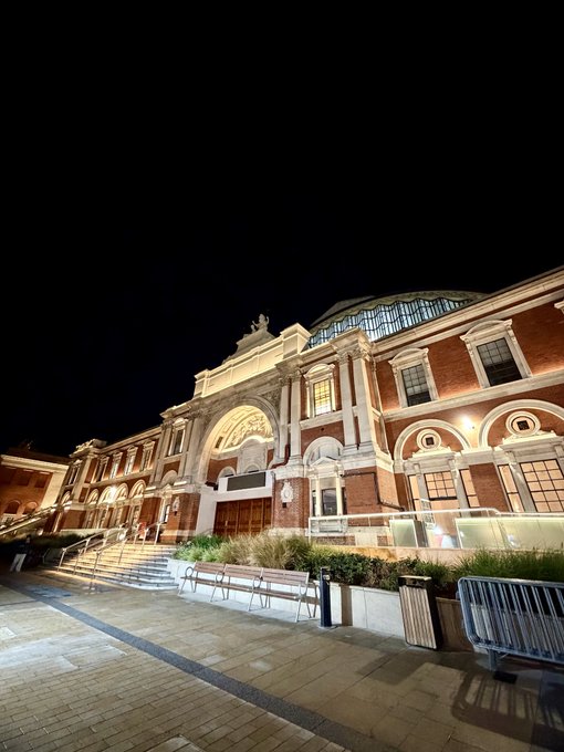 A nighttime photograph of an ornate, illuminated historic building with red brick and stone architecture, possibly a theater or museum, with steps leading to its entrance.