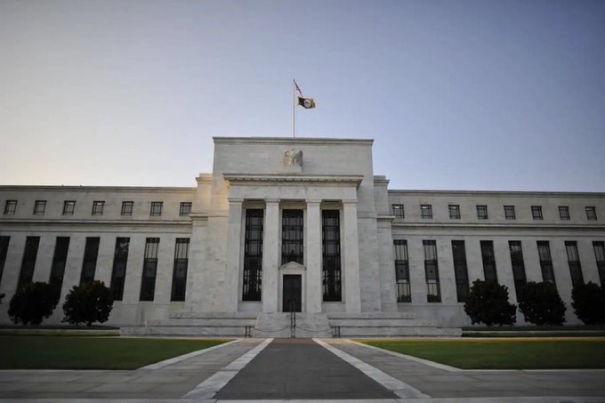 The image shows the Federal Reserve building in Washington D.C. with its flag flying above the neoclassical marble façade featuring columns and the seal above the entrance. This relates to the breaking news as the Federal Reserve, shown in the image, is the institution that would be prohibited from issuing a Central Bank Digital Currency (CBDC) until 2030 under the newly passed bill.