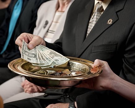 A person in a dark suit passes a silver collection plate filled with coins while another person places dollar bills into it, likely during a church service.