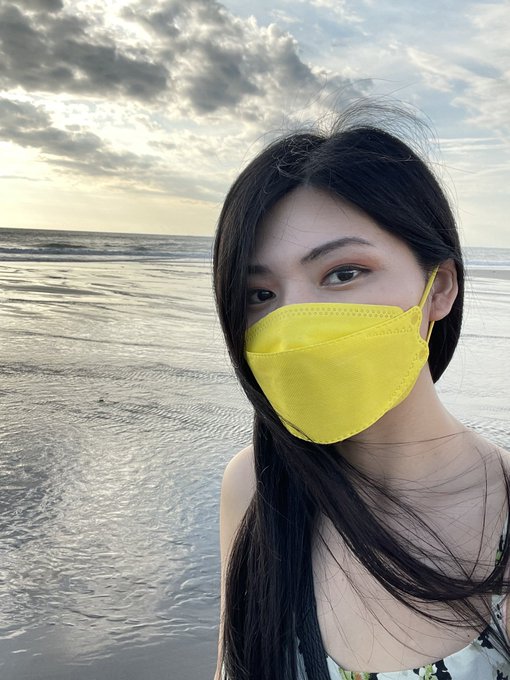 A young woman with long dark hair poses on a beach at sunset wearing a bright yellow face mask, capturing the seaside atmosphere of Bali. The image evokes the tropical setting referenced in the post about Coinfest Bali, suggesting she's at the beach location during the event.