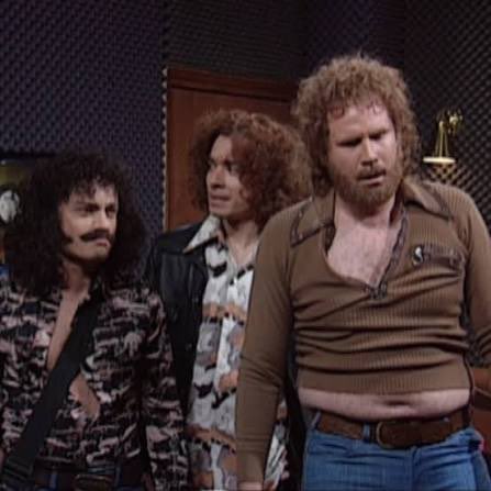Three men with 1970s-style hair and shirts stand in a dimly lit room, appearing to be from a sketch or comedy show.