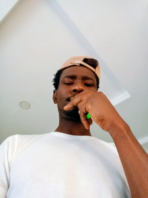 A low-angle selfie of a man in a white t-shirt wearing a backwards tan cap, with his hand near his mouth and a green ring visible on his finger.