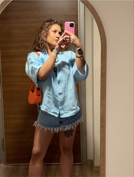 A mirror selfie of a person with curly hair wearing a light blue short-sleeved button-up shirt and a frayed denim mini skirt, holding a phone with a pink case and carrying an orange crossbody bag.