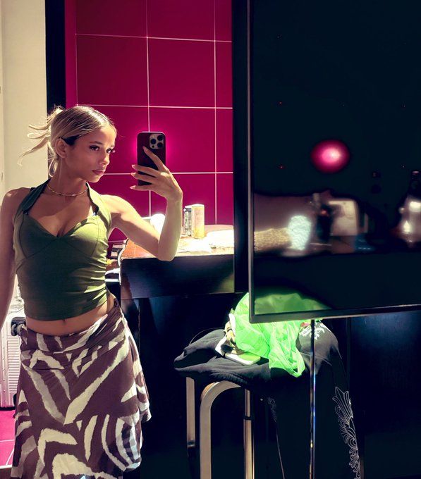 A blonde woman wearing an olive green halter top and zebra-print wrap skirt takes a mirror selfie with her phone in a room with red tile walls.