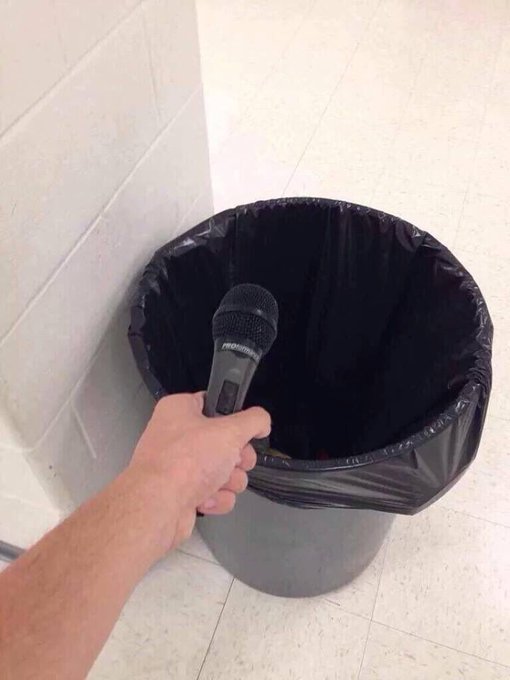 A hand holding a black microphone over a black plastic-lined trash can against a white tiled background.