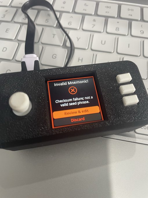 A photo of a black hardware cryptocurrency wallet device resting on a white keyboard, its screen displaying a red error message 'Invalid Mnemonic! Checksum failure: not a valid seed phrase' with 'Review & edit' and 'Discard' options.