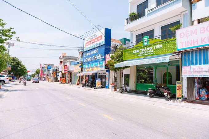 A sunny street view in a Vietnamese town featuring storefronts, including a pharmacy named 'Nhà Thuốc Long Châu' and a money transfer service called 'Kiều Hối Đông Á'.