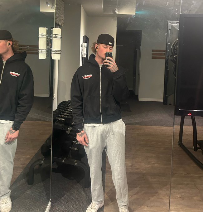 A mirror selfie of a person wearing a black "Come In" hoodie, grey sweatpants, and a black backwards baseball cap, standing in a gym.