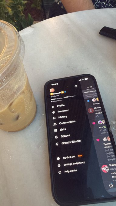 A phone displaying the Twitter/X profile menu rests on a marble table next to an iced coffee, showing notifications, options like "Premium+" and "Creator Studio," and the handle "@ballerxbt" in the search bar. The caption "Chill boi w/ @ballerxbt" complements the relaxed café scene, suggesting the user is browsing the crypto influencer's profile while enjoying a drink.