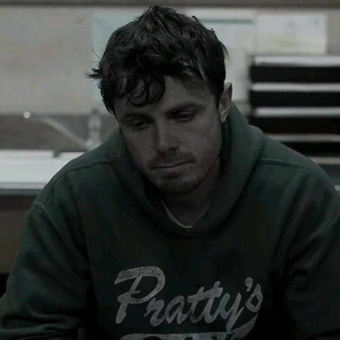 A somber portrait of a young man with messy dark hair wearing a dark green sweatshirt with "Pratty's" printed on it, looking downward in what appears to be a dimly lit room.
