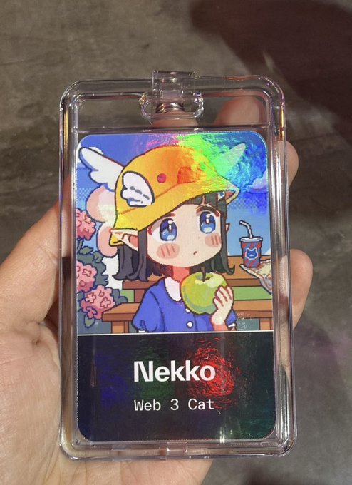 A hand holding a holographic acrylic ID badge featuring an anime cat-girl character wearing a yellow cap and holding an apple, labeled 'Nekko - Web 3 Cat.'