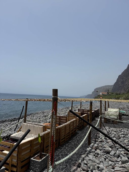 A peaceful seaside view features a rocky beach, wooden docks, and distant cliffs under a clear blue sky. The tranquil coastal setting offers a refreshing "GM" moment to start the day.
