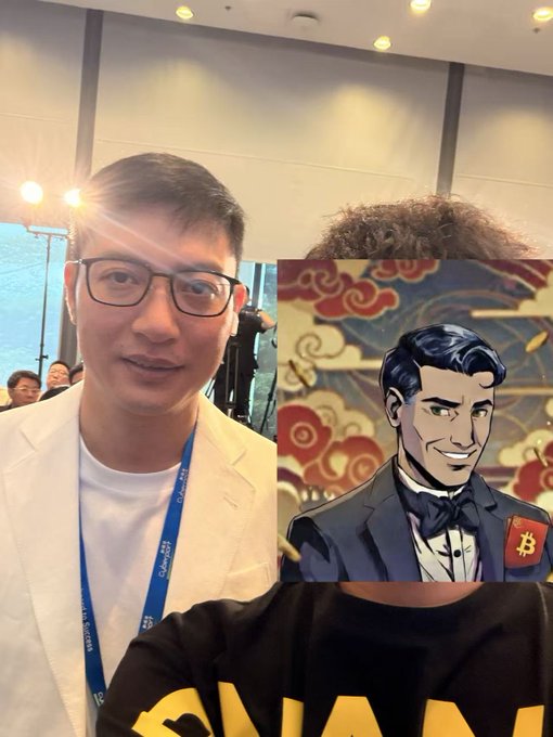 A man wearing glasses and a white blazer with a lanyard poses next to a cartoon illustration of a tuxedoed character with a Bitcoin insignia, set against a red ornamental background.