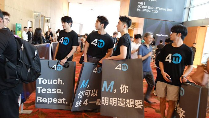 A group of young men wearing black "4m" branded t-shirts hold signs reading "Touch. Tease. Repeat." and Chinese phrases at what appears to be a conference, with a banner above indicating "LEVEL 5 DKK MAIN STAGE: BIT DIGITAL INSTITUTIONAL STAGE" and "MetaPersona."

The image shows promotional activity for @4Mexchange, the cryptocurrency exchange referenced in the post, with staff distributing marketing materials at the Bit Digital Institutional Stage, visually representing the exchange's presence and entry into the market during a downturn as discussed.