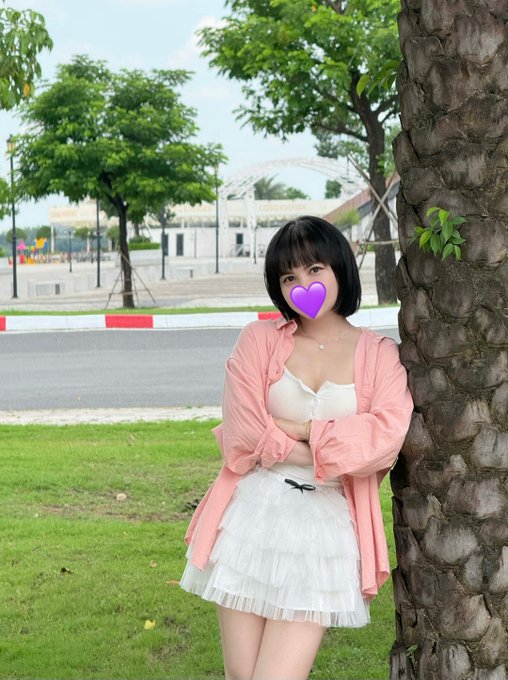 A woman with short black hair poses in a pink shirt and white skirt by a tree, with a purple heart emoji covering her face, while in the background a "CONG HOA" sign is visible on a building across the street. The casual outdoor pose mirrors the post's laid-back, grind-mood vibe of still pursuing passions and holding NFTs without profit in mind.