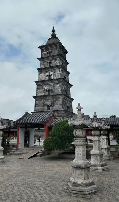 A tall, multi-tiered stone pagoda rises behind ornate stone lanterns and traditional Chinese temple architecture with curved roofs and red accents.