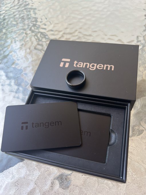 A black Tangem wallet box sits open on a stone surface, displaying a smart ring and two branded cards with the "tangem" logo visible, illustrating the brand's hardware wallet product line mentioned in the post.
